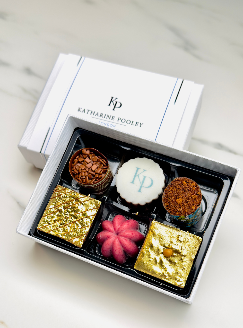 An open white Katharine Pooley gift box containing assorted gourmet chocolates from Giuliana Pimenta photographed on a light marble surface. The box lid is placed behind the open tray, showing the KP brand name and logo.