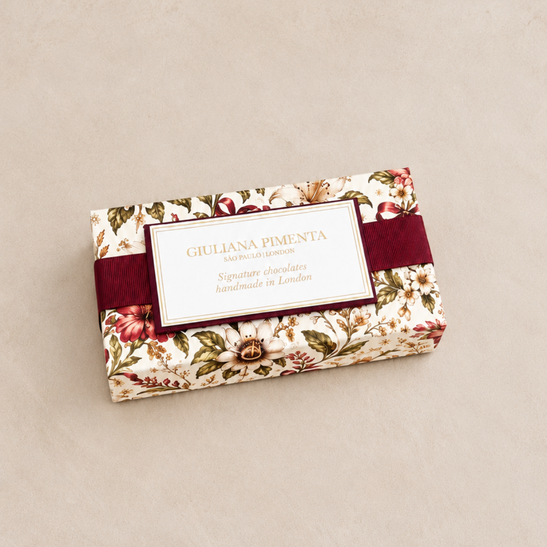 A rectangular chocolate box wrapped in cream floral paper with red and green botanical print, tied with a dark red ribbon and featuring a white label that reads 'GIULIANA PIMENTA SIGNATURE CHOCOLATES handmade in London'.