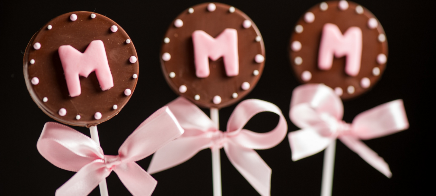 Three chocolate lollipops on a black background, each stamped with a pink
letter M framed by a border of white dots, finished with pink satin bows
