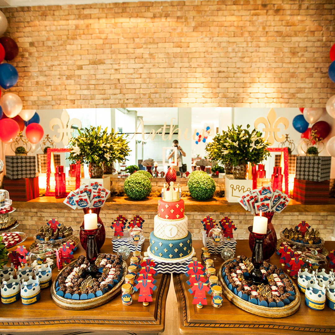 Dessert table for a first birthday party in red, blue and gold, with a
three tier cake marked with the number one, fleur-de-lis decorations,
balloons, candles and gold trays of brigadeiros and chocolates