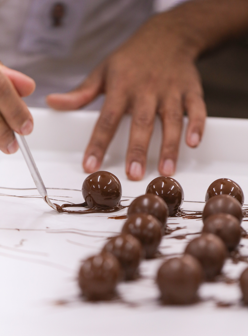 Chocolate-covered spheres being dipped with chocolate on a white surface, with a person's hands visible in the background at Giuliana Pimenta's London luxury chocolate atelier hand making the bonbons