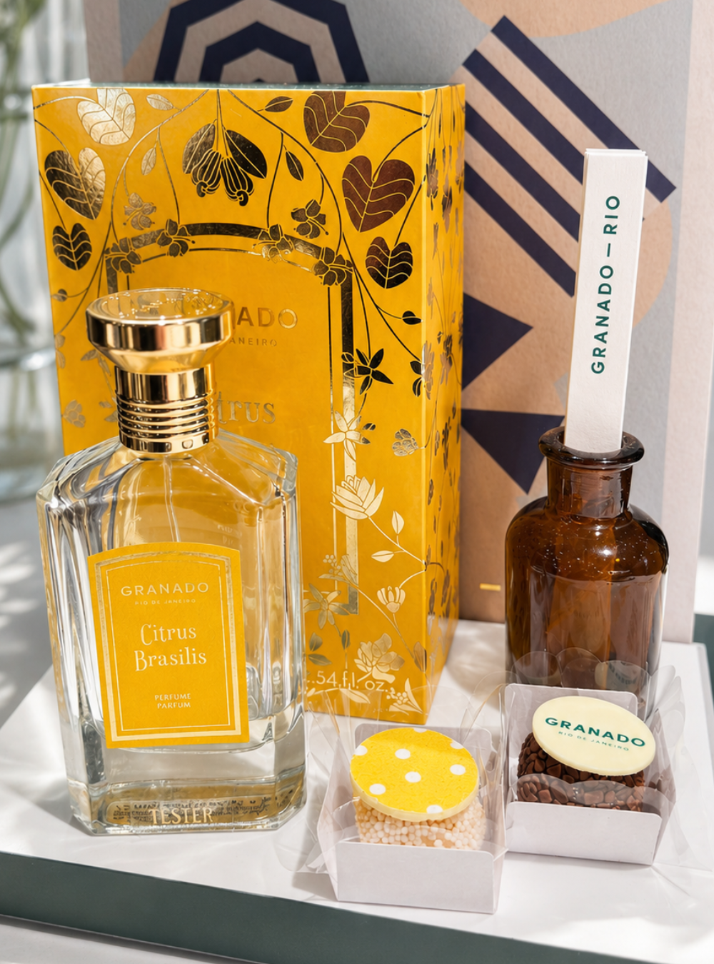 Granado gift set featuring a Citrus Brasil liqueur bottle, a yellow patterned box, a brown diffuser bottle with a white wand, and two small personalised brigadeiros with chocolate disc displaying Granado's brand and the packaging pattern, handmade by Giuliana Pimenta in our London luxury chocolate atelier.