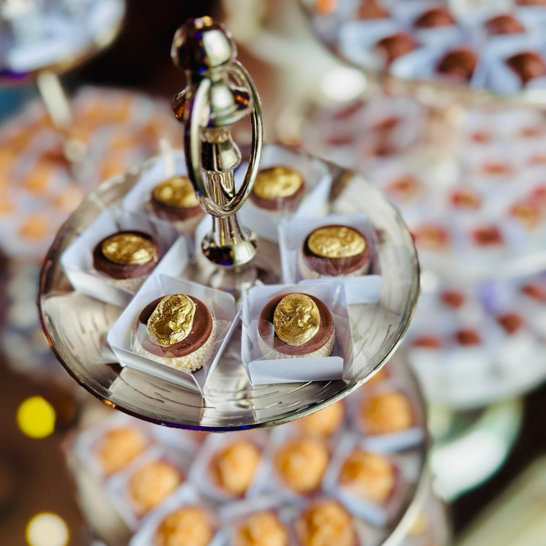 A close-up of assorted chocolates arranged on a clear glass tiered stand with a silver central finial. Several chocolates are topped with shiny gold leaf, and the background shows more chocolates in soft focus.