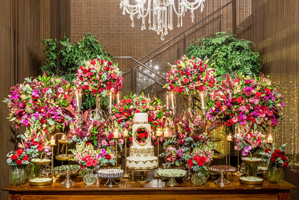 Luxury dessert table for a birthday celebration, with a tiered white and gold cake, tall red and pink floral arrangements, lit candelabras and gold stands of brigadeiros and handmade chocolates