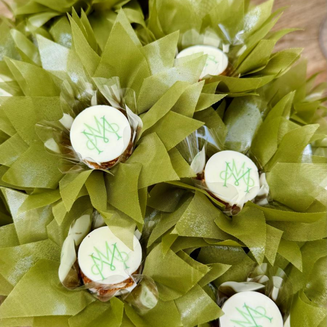 A close-up of several chocolates arranged together on a luxury sweet table for a Wedding at Hampton Court House, each topped with a white chocolate piece printed with the couple's green monogram lettering, surrounded by olive-green flower-shaped chocolate paper case. 