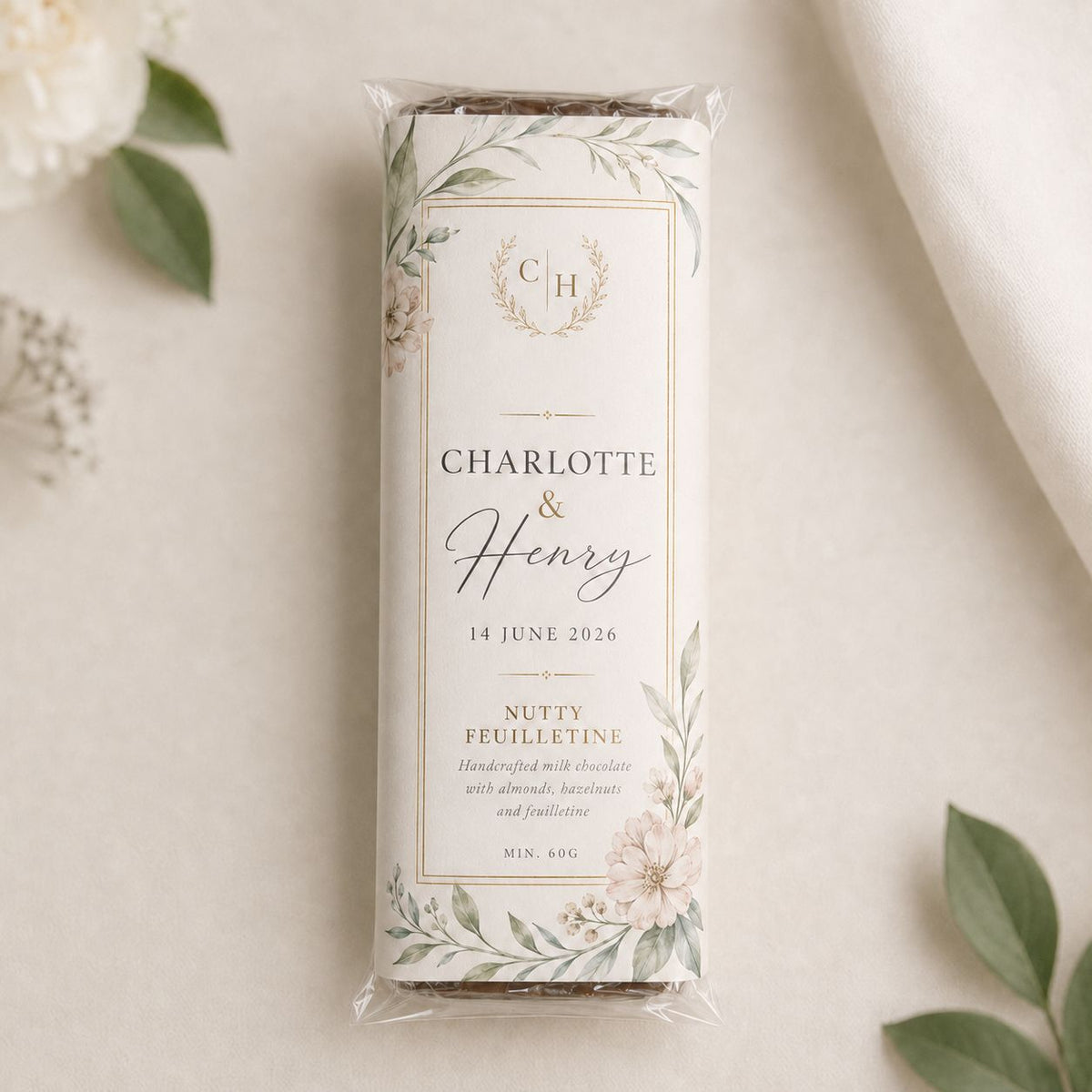 A personalised chocolate bar wrapped in a bespoke paper sleeve, featuring a floral label with the names 'Charlotte & Henry' and the date '14 June 2026'. The bar is photographed on a light beige textured surface with soft white flowers and green leaves in the corners.
