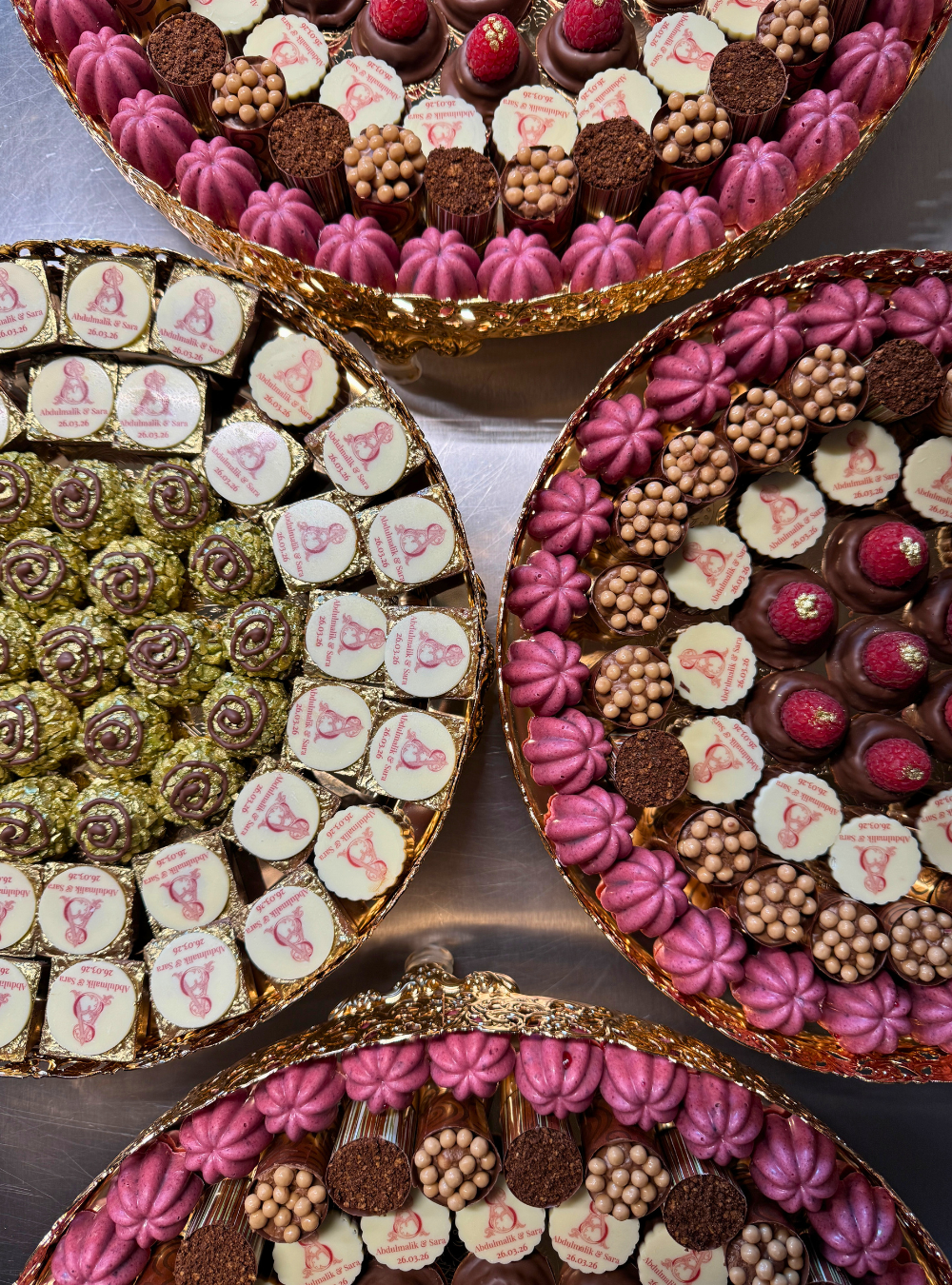 Four gold trays of personalised chocolates for an Arabic wedding in London, each piece printed with the couple's monogram and wedding date, arranged in berry and rose tones with chocolate-dipped raspberries finished in gold leaf
