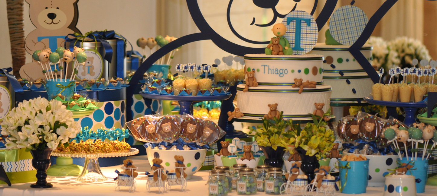 Chocolate table for a first birthday party, teddy bear themed in navy blue and green, with a tiered cake iced with the name Thiago, bear-faced cake pops and chocolate lollipops, brigadeiros in cups and jars of sweets